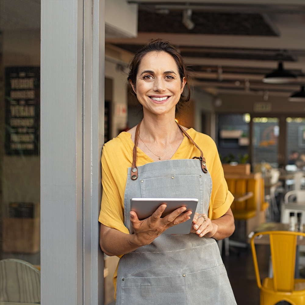 A small business owner standing outside of her store holding an ipad and smiling.
