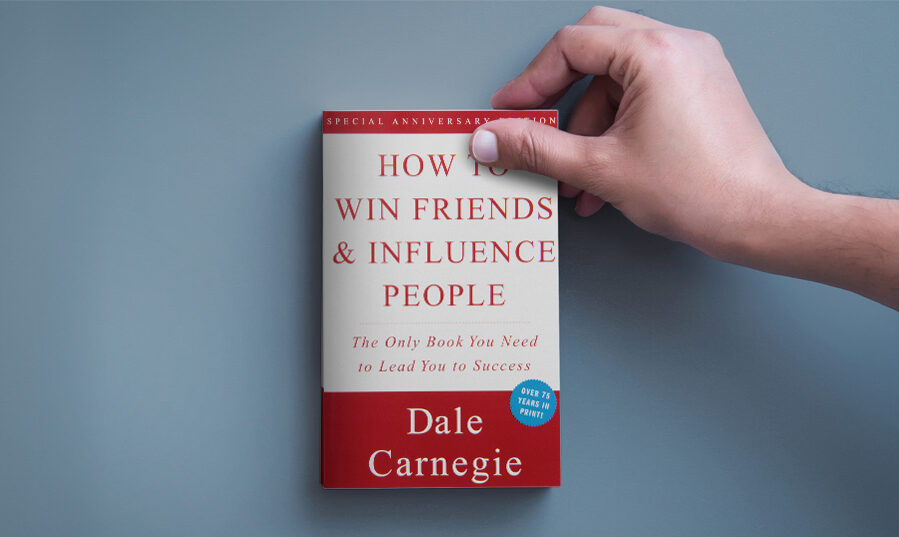 A person holding the corner of a copy of How to Win Friends and Influence People that's sitting on a desk