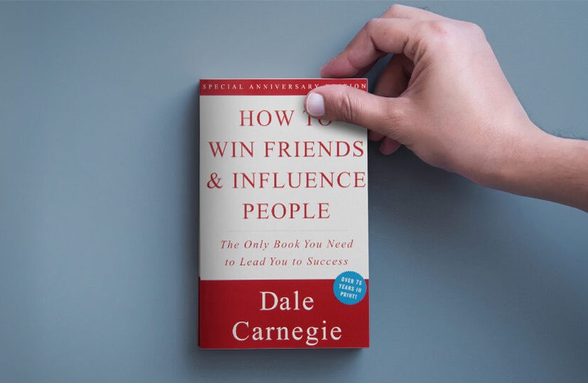 A person holding the corner of a copy of How to Win Friends and Influence People that's sitting on a desk